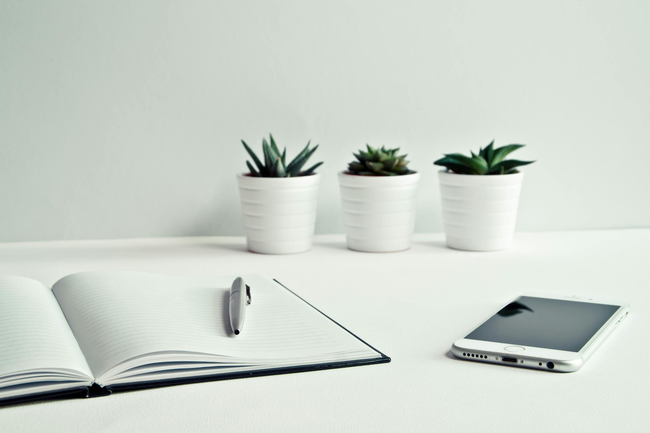 A clean, minimalist workspace featuring a smartphone, open notepad, pen, and potted succulents.
